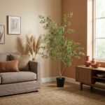 Living room scene with a gray sofa, orange and sage cushions, a tall leafy plant in a black pot, and a wooden sideboard by a window.