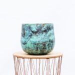 Turquoise ceramic planter with a weathered glaze sitting on a round wooden tabletop supported by copper wire legs.