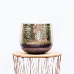 Textured, multicolored glass vase with a gradient from green to purple sits on a round wooden pedestal atop a copper-wire table (interior decor).