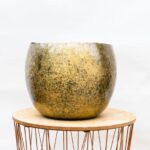 A weathered ceramic planter with a gold-brown glaze sits on a round wooden table with a copper wire base outside a white background.