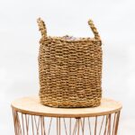 Woven wicker basket with two handles atop a round wooden table on a light background.