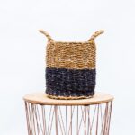 Two-tone woven basket (natural and dark blue) on a round wooden table with thin metal legs, photographed against a light background.