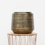Bronze-toned ceramic vase on a round wooden table with a copper-wire base stand.