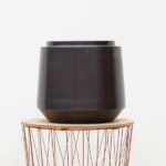 Dark matte planter sits on a round wooden table with a copper wire base/stand.