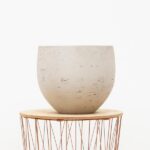 Beige ceramic bowl with a textured surface resting on a round wooden top with a copper wire stand, against a white background.