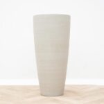 Tall cylindrical ceramic vase in light gray-beige on a wooden surface on a white background.