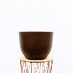 Brown ceramic planter resting on a light wooden round tray, supported by a copper wire stand against a white background.