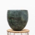 Large dark green textured ceramic planter on a round wooden table against a white background, empty without plants inside.